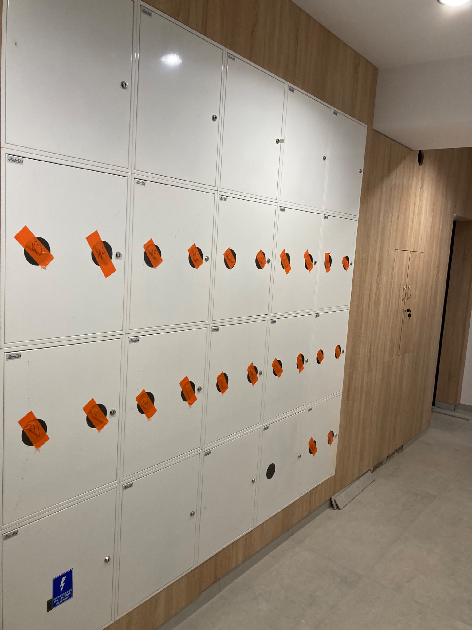 Rząd białych szafek z numerami na pomarańczowych naklejkach w Brodnicy, zamontowanych na ścianie z drewnianą okładziną.