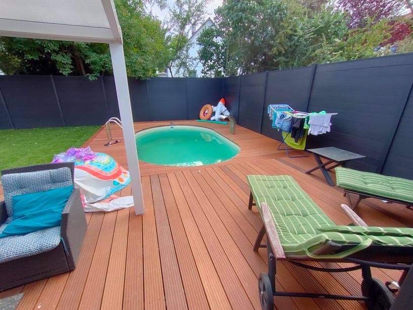 Drewniany taras z małym basenem, leżakami z zielonymi poduszkami i czarnym ogrodzeniem w tle, pranie suszy się na suszarce.