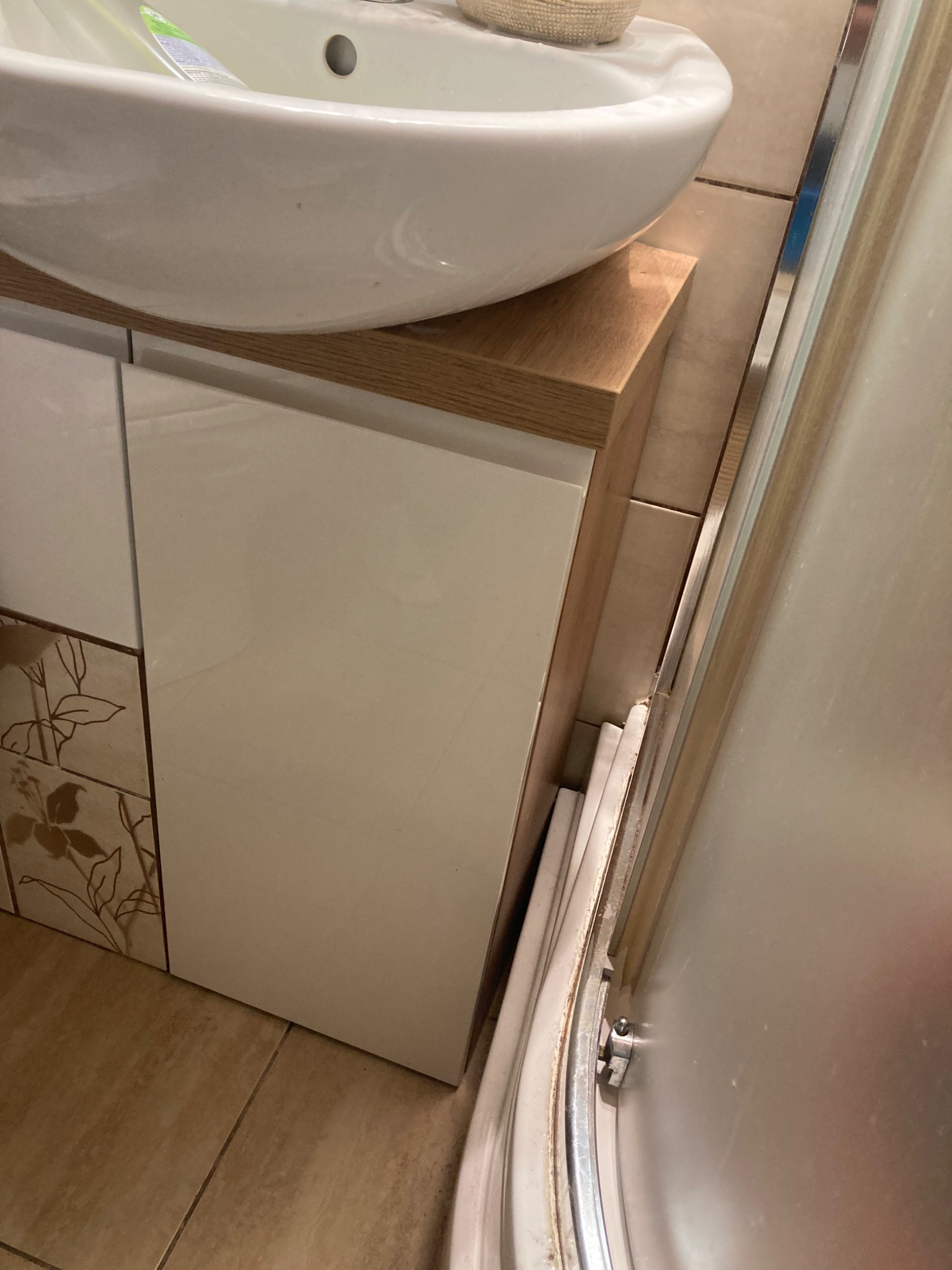 Biała umywalka nablatowa na szafce z drewnianym blatem, obok kabina prysznicowa z widocznym profilem. Dekoracyjne płytki z motywem roślinnym na ścianie.