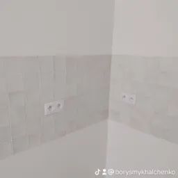 Fragment ściany wyłożonej jasnymi kwadratowymi płytkami ceramicznymi z gniazdkami elektrycznymi w Gdyni, widok z narożnika pomieszczenia.