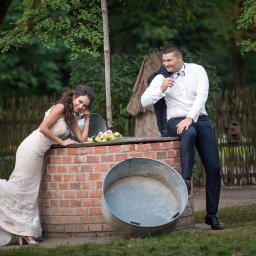 Usługi Video - Uśmiechnięta panna młoda w koronkowej sukni oparta o ceglany murek, obok siedzący pan młody w muszce i marynarce zarzuconej na ramię, w tle zieleń drzew.