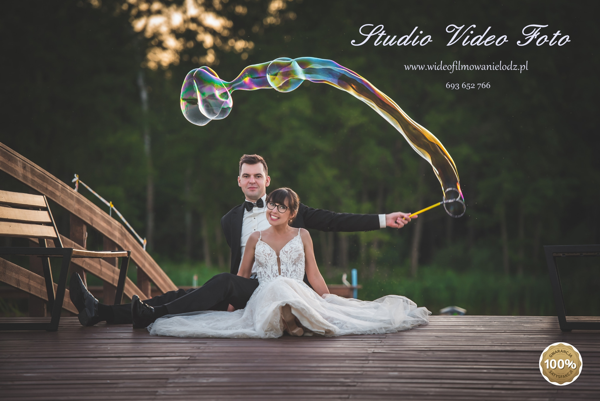 Para młoda siedzi na drewnianym pomoście, mężczyzna w smokingu puszcza duże, tęczowe bańki mydlane. W tle zielone drzewa i jezioro.