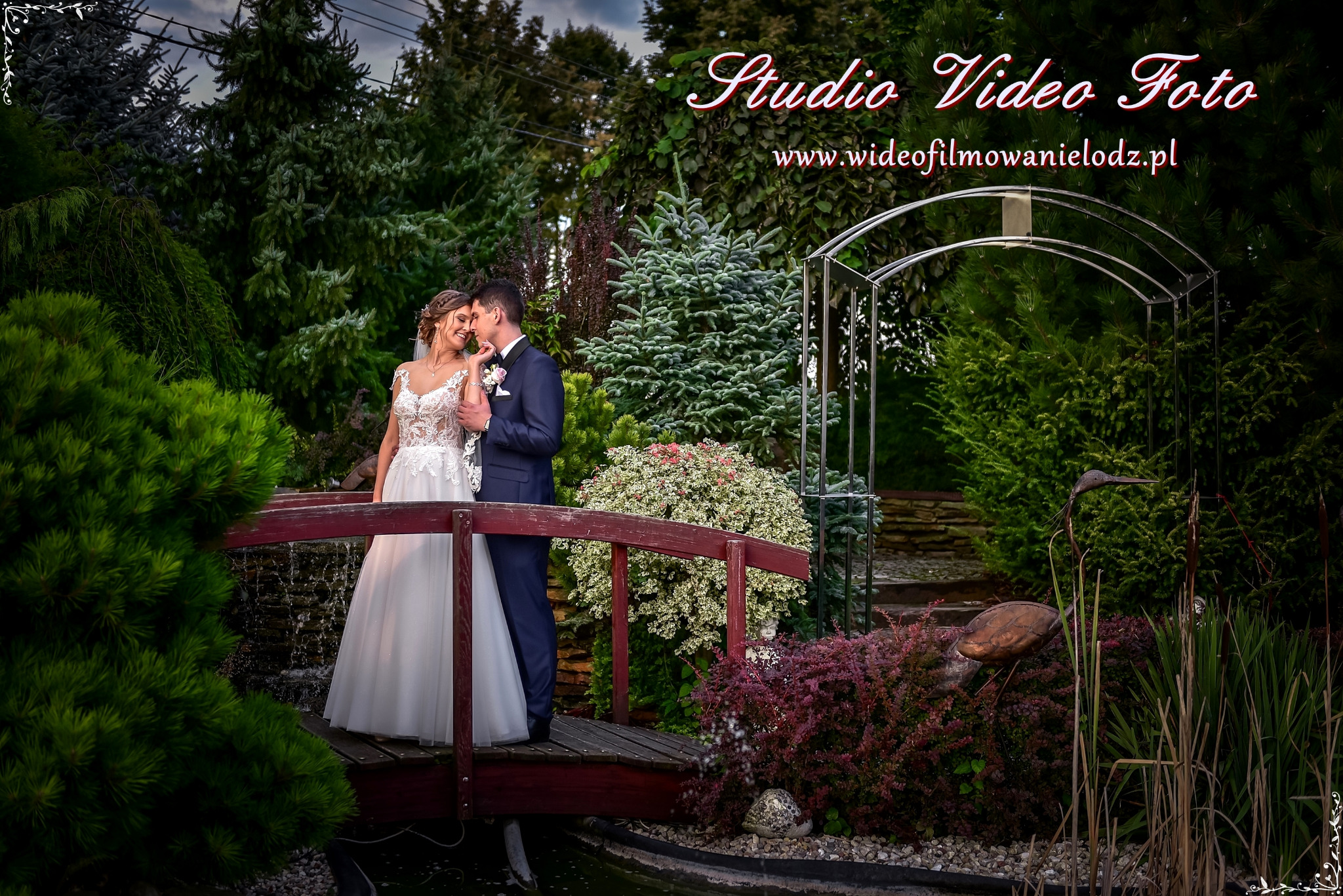 Para młoda, on w granatowym garniturze, ona w białej sukni z koronką, stoi na małym drewnianym mostku w ogrodzie, w tle zielona roślinność i dekoracyjna metalowa konstrukcja.