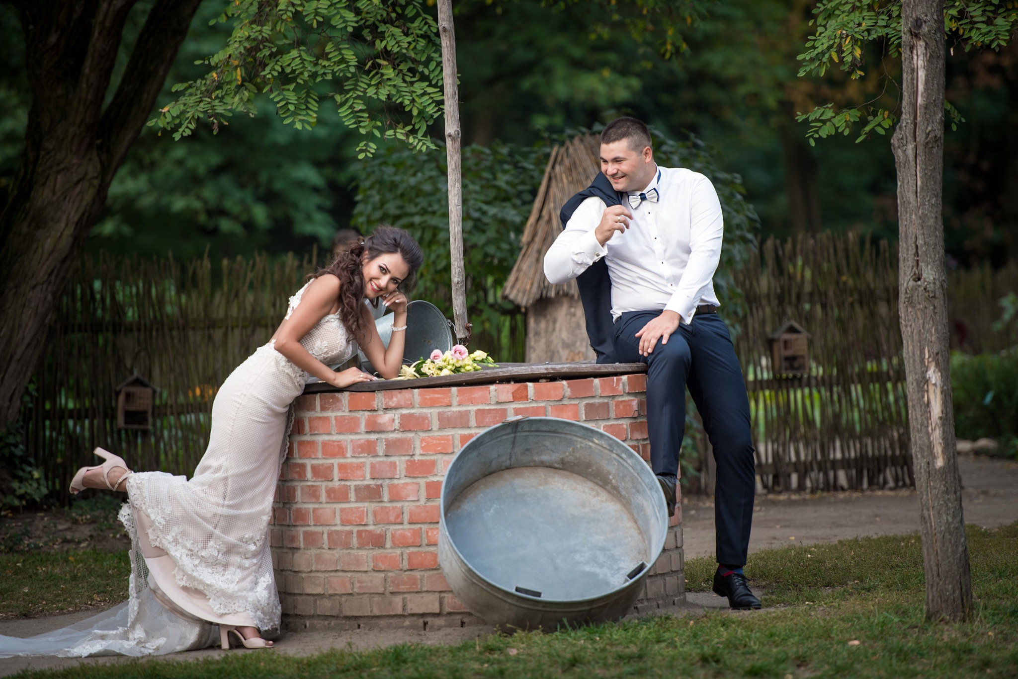 Uśmiechnięta panna młoda w koronkowej sukni oparta o ceglany murek, obok siedzący pan młody w muszce i marynarce zarzuconej na ramię, w tle zieleń drzew.