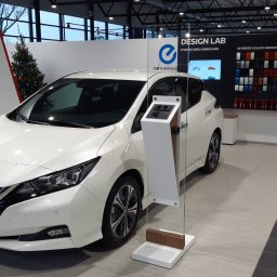 RTS sp. z o.o. - Biały samochód elektryczny Nissan Leaf w salonie samochodowym, obok panel z próbkami kolorów nadwozia i choinka w tle.