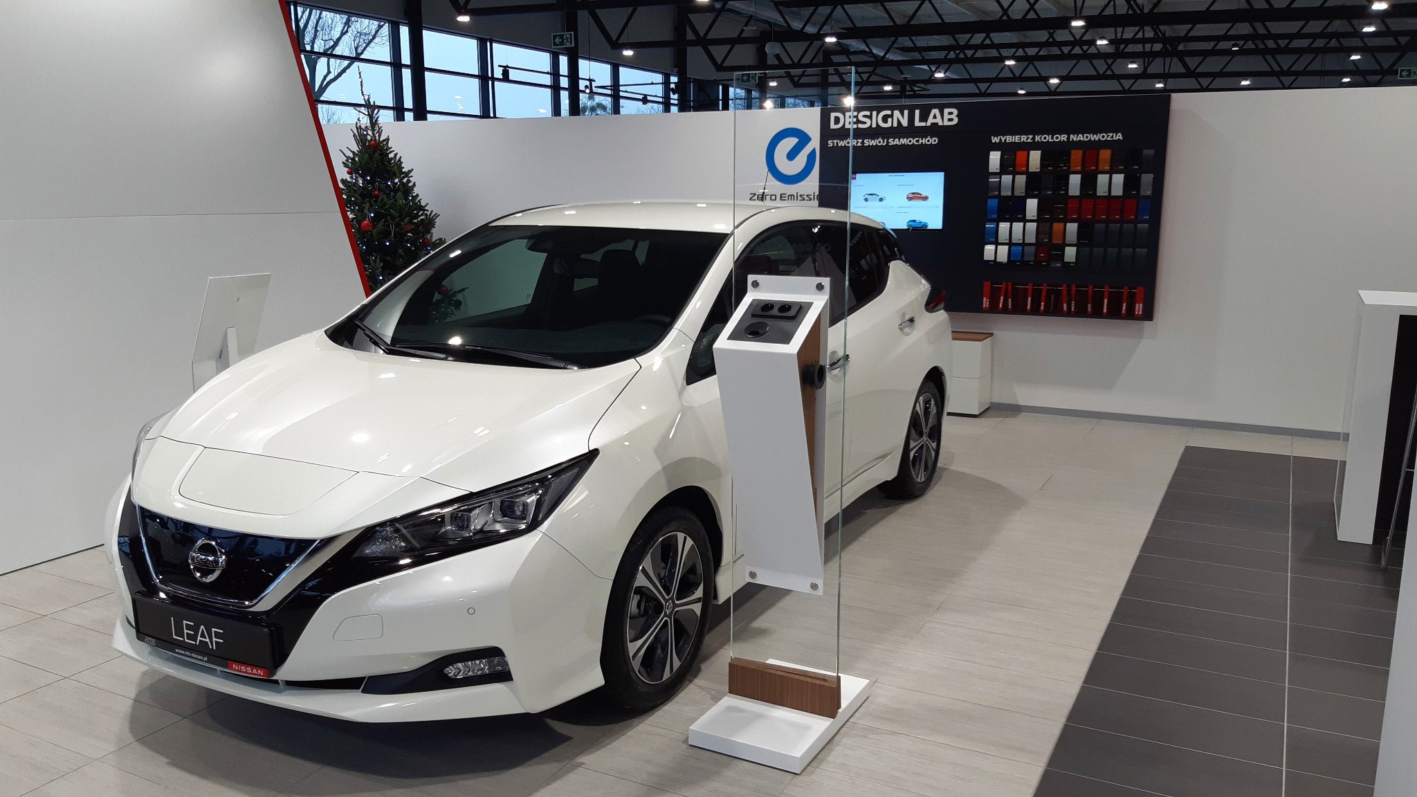Biały samochód elektryczny Nissan Leaf w salonie samochodowym, obok panel z próbkami kolorów nadwozia i choinka w tle.
