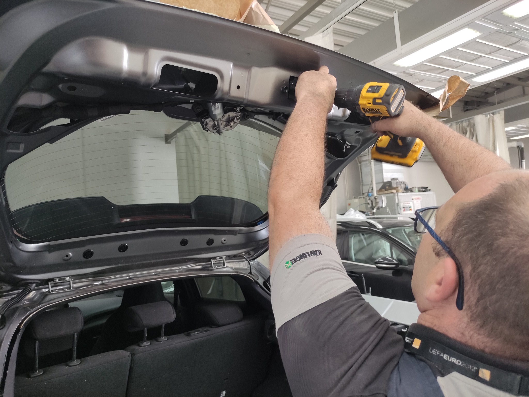 Mechanik z wiertarką DeWalt naprawia klapę bagażnika szarego samochodu w warsztacie.