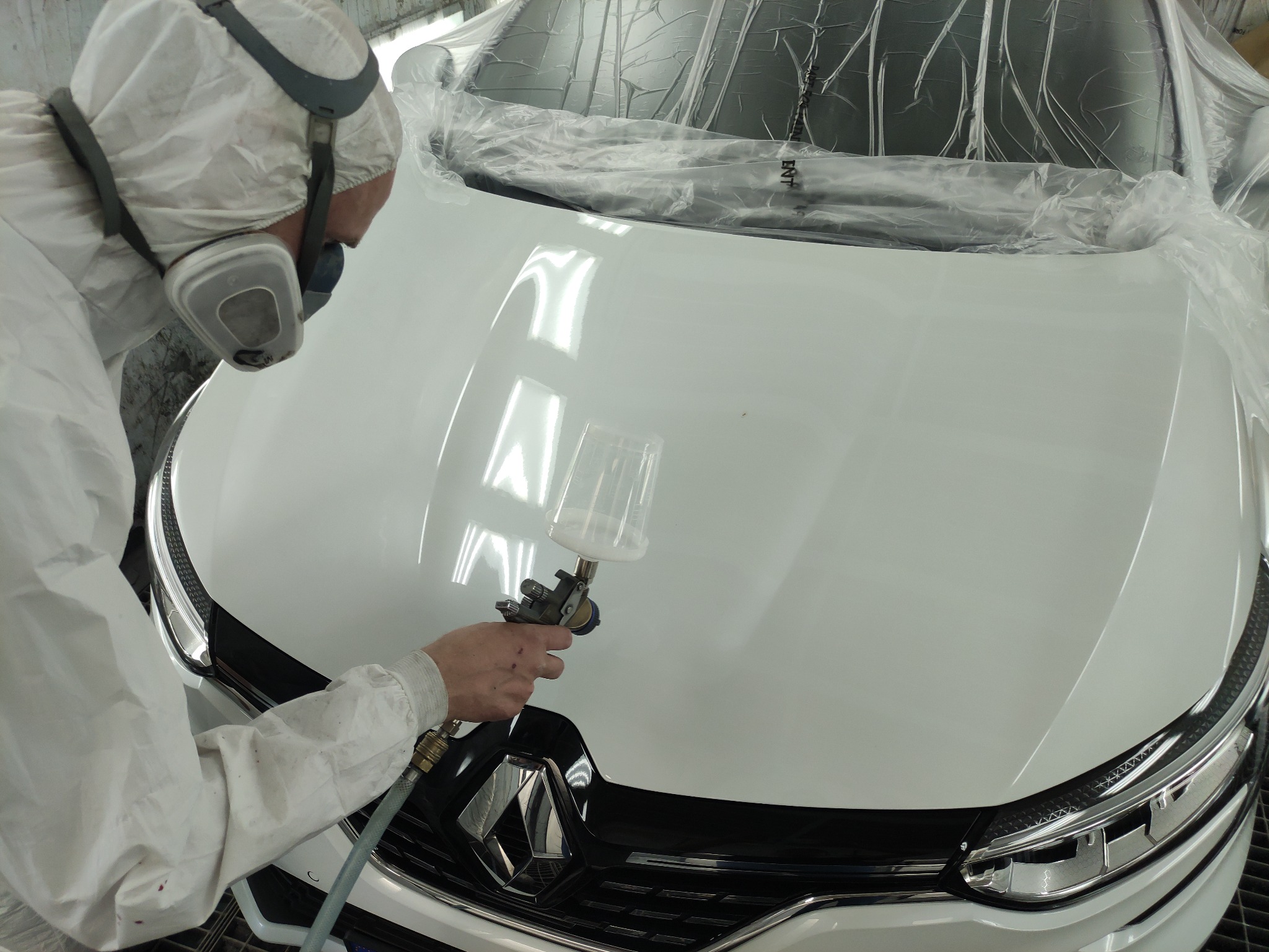 Pracownik w kombinezonie ochronnym lakieruje biały samochód pistoletem natryskowym, przednia maska i reflektory zabezpieczone folią.