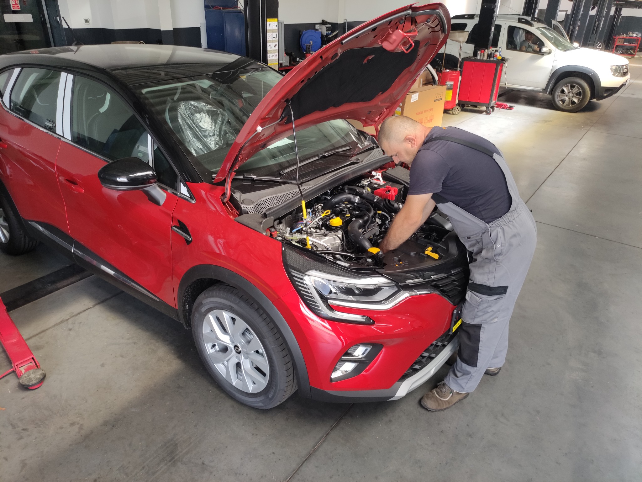 Mechanik w szarym kombinezonie pochyla się nad otwartą maską czerwonego samochodu w warsztacie, widoczny silnik i elementy instalacji.