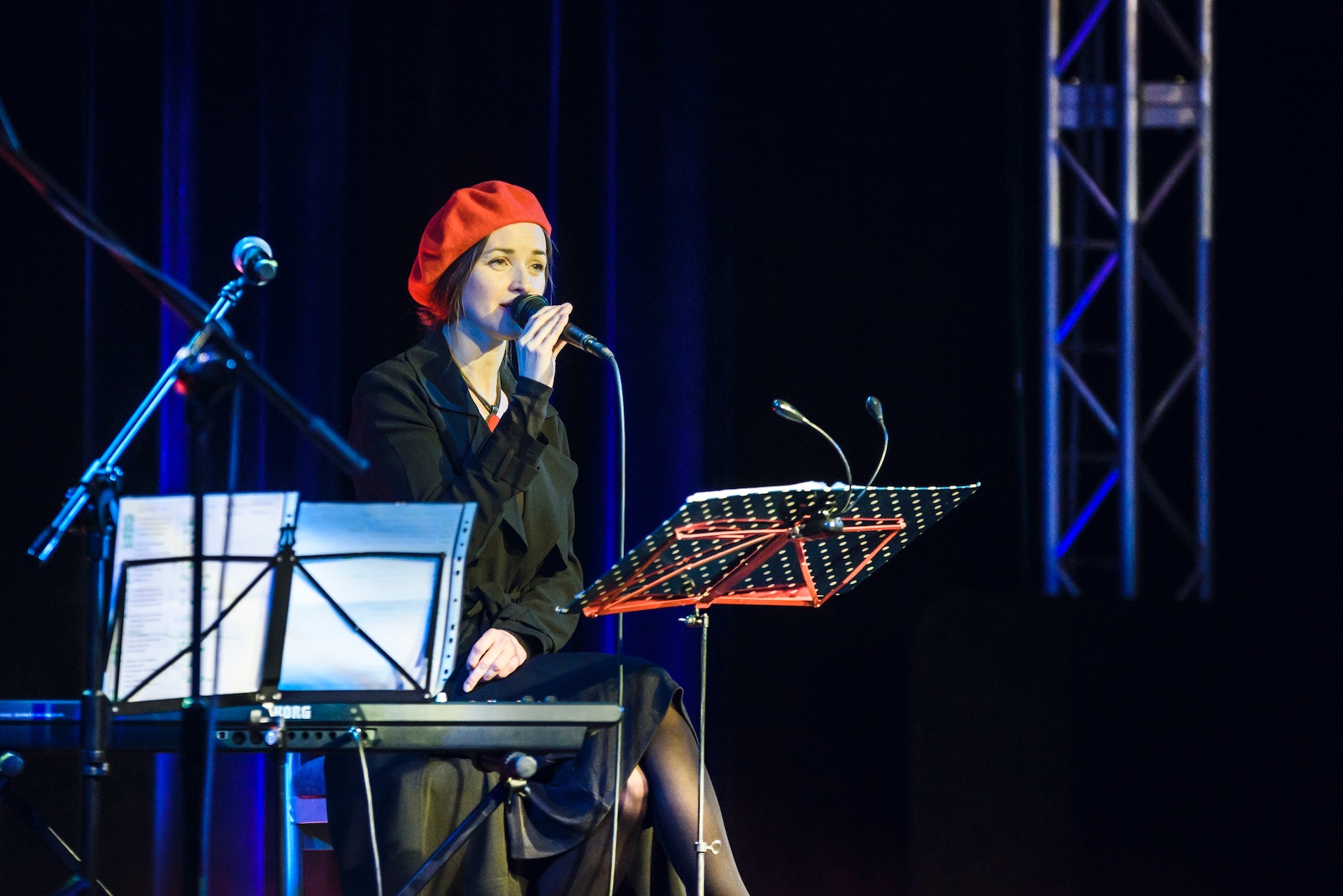 Kobieta w czerwonym berecie śpiewa do mikrofonu, siedząc przy keyboardzie Korg na scenie z ciemnoniebieskimi zasłonami w tle i metalową konstrukcją oświetleniową po prawej stronie.