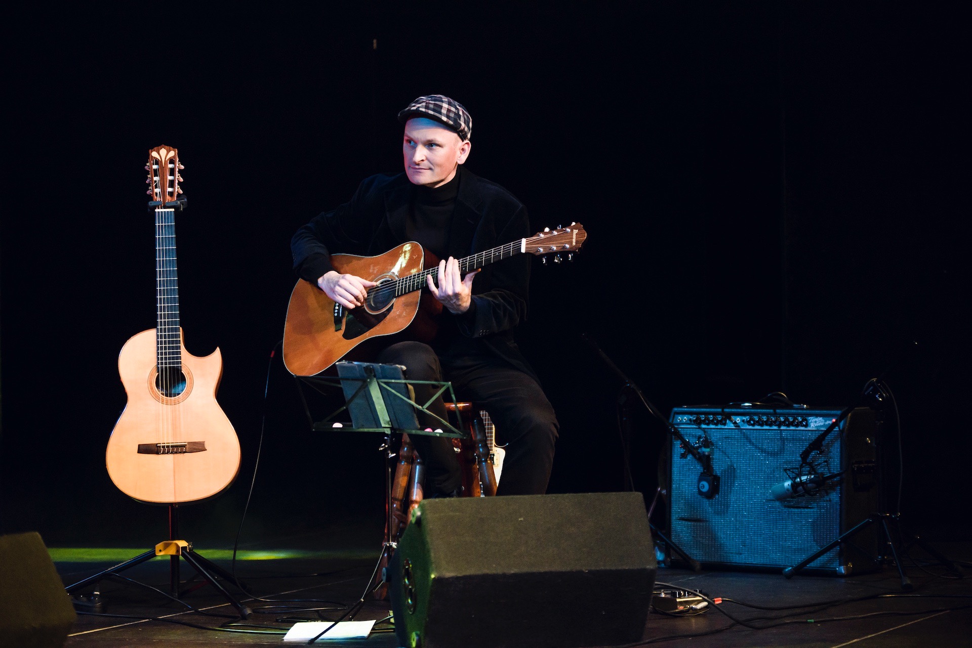 Gitarzysta w czarnym stroju i czapce gra na akustycznej gitarze na scenie, obok stoi klasyczna gitara, widoczny wzmacniacz i mikrofony.