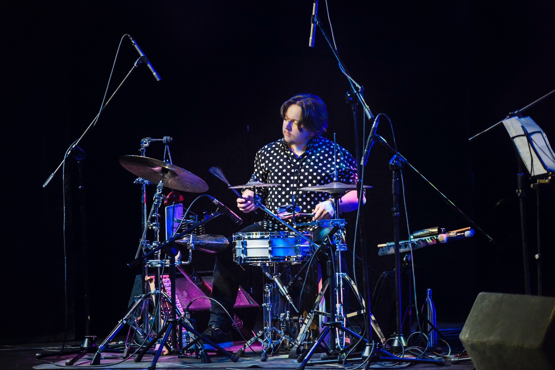 Perkusista w koszuli w grochy gra na perkusji na scenie z licznymi mikrofonami i statywami.