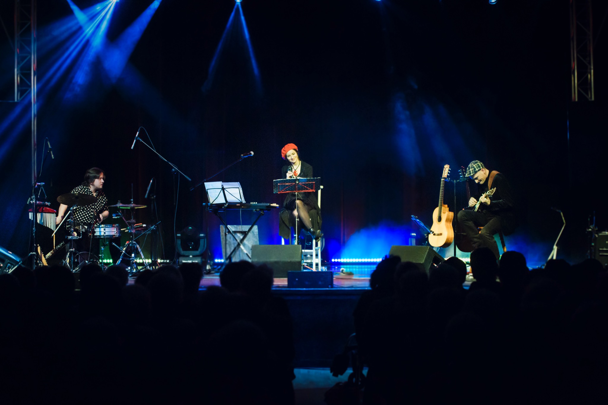 Scena z koncertu: perkusista w koszuli w kropki, wokalistka w czerwonym berecie, gitarzysta w czapce, niebieskie oświetlenie i publiczność w tle.