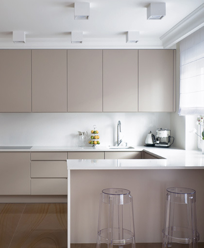 Nowoczesna kuchnia z beżowymi szafkami i białym blatem, przezroczyste krzesła barowe, oświetlenie punktowe i drewniana podłoga.