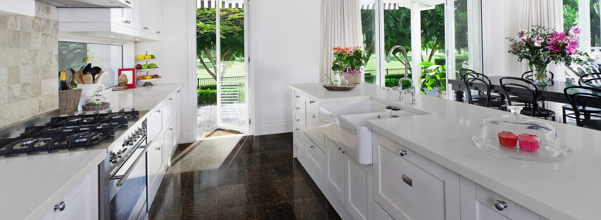 Jasny blat kuchenny z białymi szafkami, dużym zlewem i widokiem na ogród przez otwarte drzwi balkonowe, na blacie dekoracje i babeczki pod kloszem.