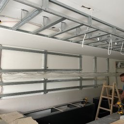 ALUX firma budowlana - Montaż metalowej konstrukcji sufitu podwieszanego z płyt kartonowo-gipsowych, widoczne przewody elektryczne, częściowo zamontowane płyty, drabina i narzędzia w tle.