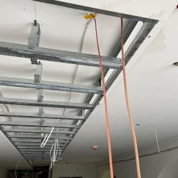 ALUX firma budowlana - Metalowa konstrukcja sufitu podwieszanego z widocznymi przewodami elektrycznymi, mocowana do białego sufitu z plamami wilgoci.