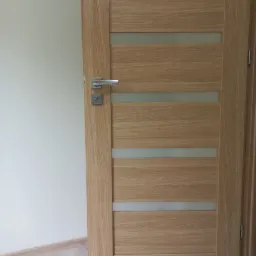 Otwarta, drewniana, wewnętrzna drzwi z czterema poziomymi, matowymi szybami, srebrna klamka i zamek, widoczna podłoga z jasnych płytek i fragment ściany w neutralnym kolorze.