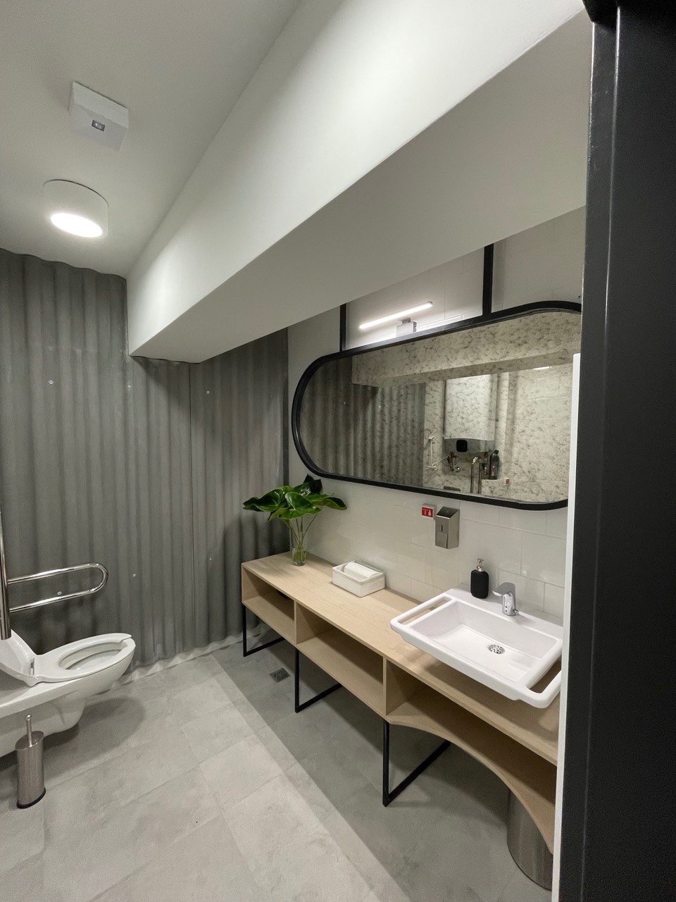 Nowoczesna łazienka z toaletą dla niepełnosprawnych, umywalką na drewnianym blacie z metalowymi nogami, lustrem w czarnej ramie i szarą, teksturowaną ścianą.