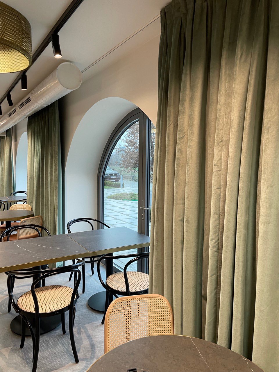 Wnętrze kawiarni lub restauracji z ciemnymi stołami, krzesłami z rattanowym siedziskiem, oliwkowymi zasłonami i łukowymi oknami wychodzącymi na zewnątrz.