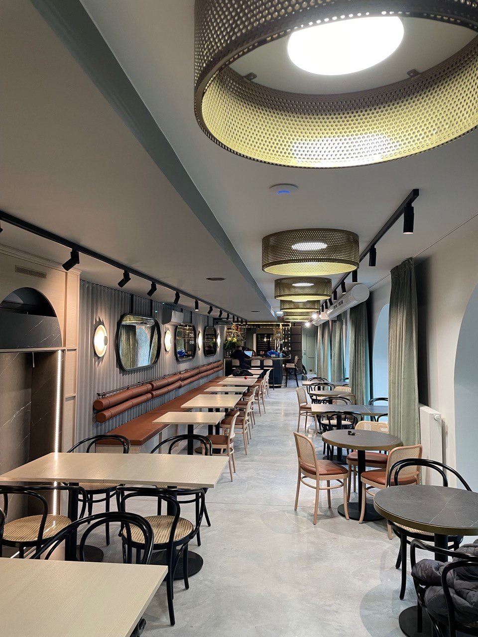 Wnętrze restauracji z rzędem stolików i krzeseł, oświetlonych wiszącymi lampami z ażurowymi kloszami, z widoczną ścianą z dekoracyjnymi lustrami i tapicerowaną ławą.