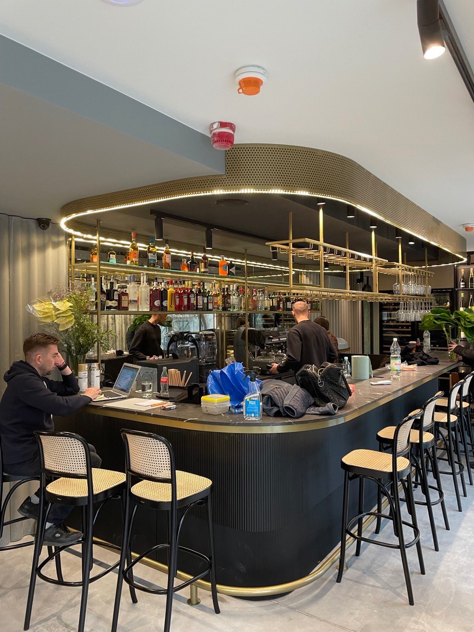 Wnętrze baru z czarnym, marmurowym blatem, złotymi akcentami i rzędem krzeseł barowych z rattanowym siedziskiem, oświetlonych dekoracyjnym sufitem z podświetleniem LED.