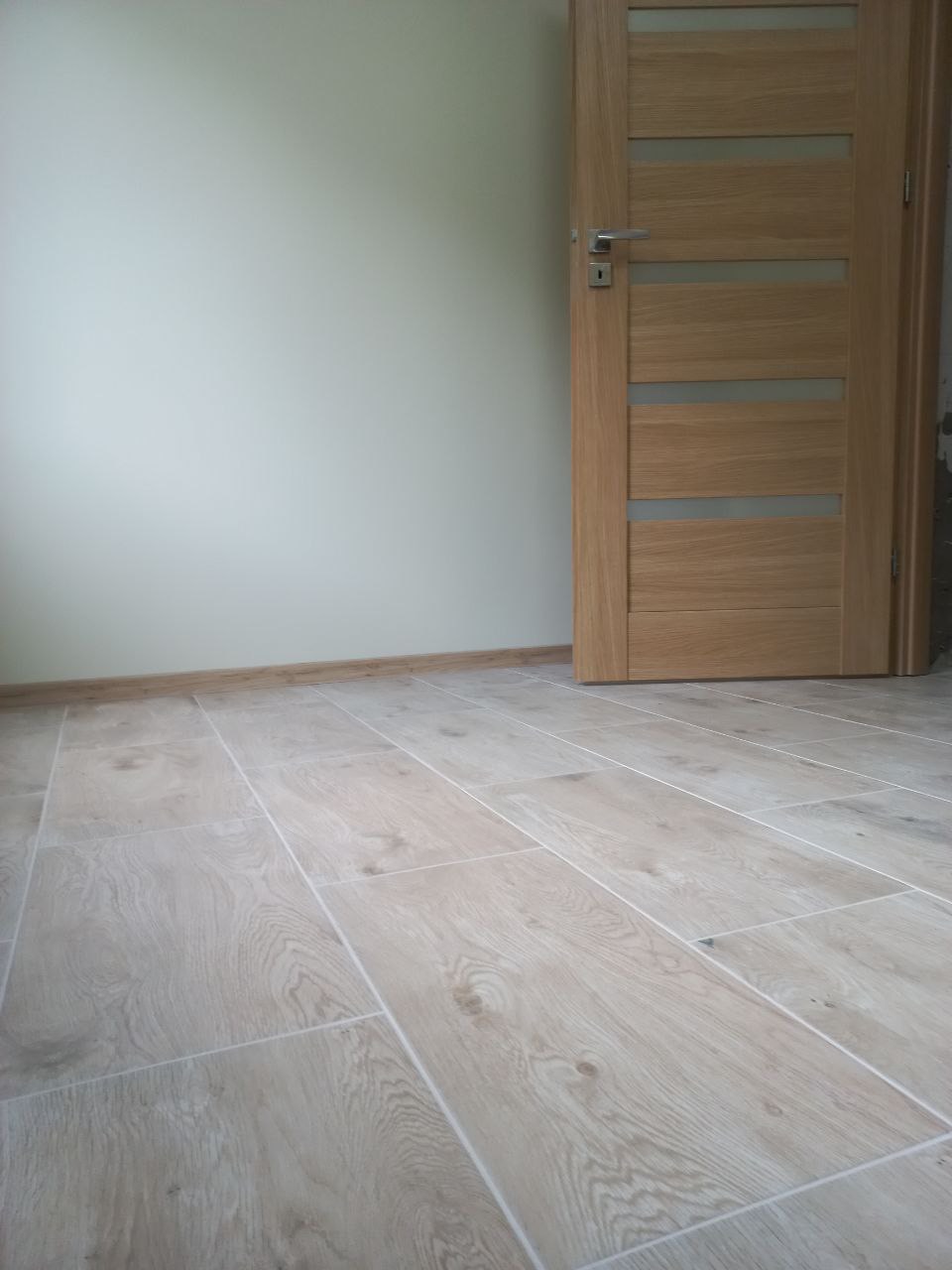 Jasne, drewnopodobne płytki podłogowe ułożone w prostokątnym wzorze, widoczne przy ścianie i drewnianych drzwiach z przeszkleniami.