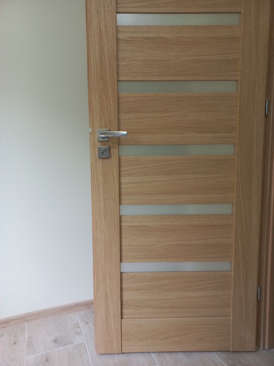 Otwarta, drewniana, wewnętrzna drzwi z czterema poziomymi, matowymi szybami, srebrna klamka i zamek, widoczna podłoga z jasnych płytek i fragment ściany w neutralnym kolorze.