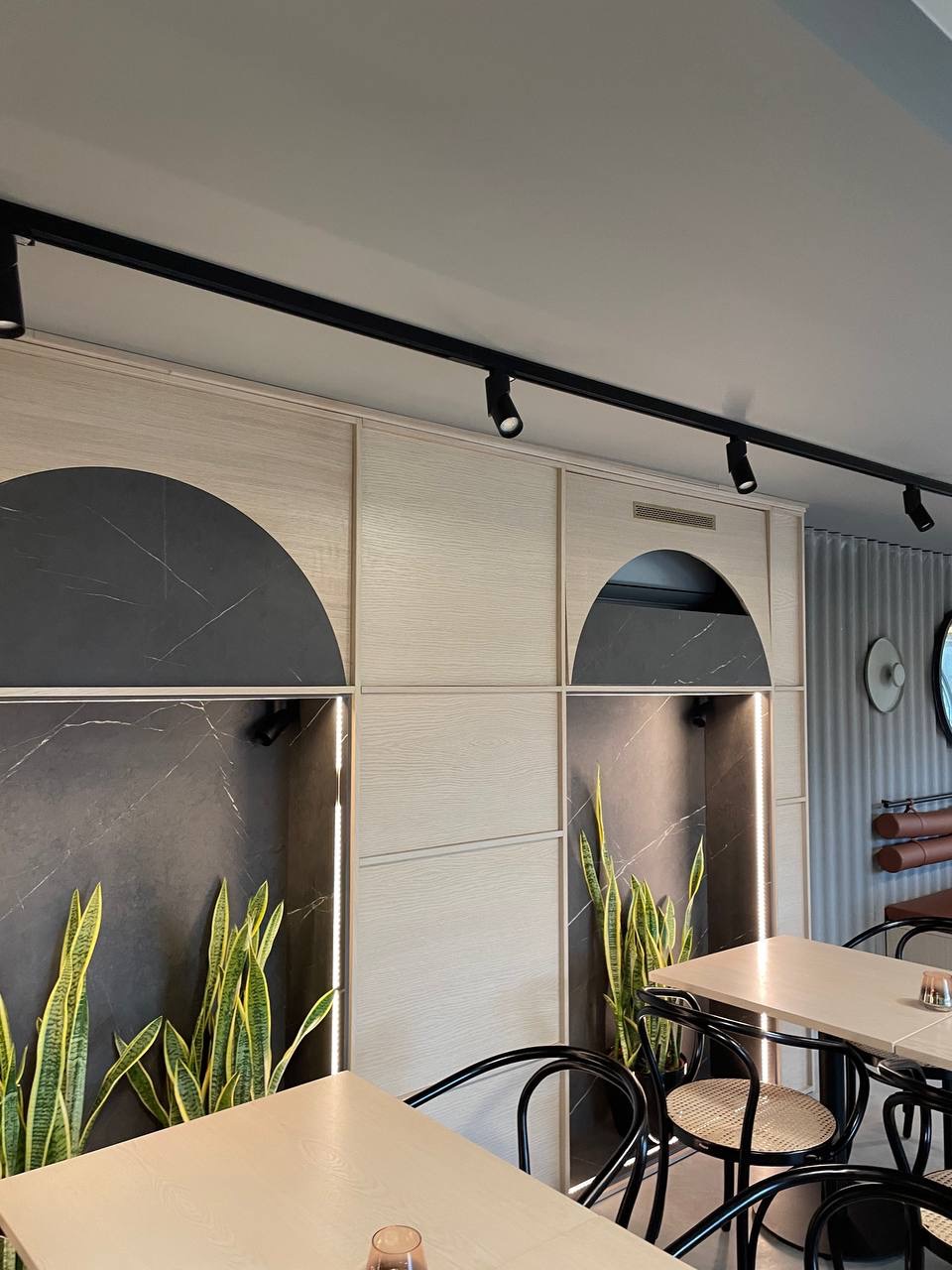 Eleganckie wykończenie wnętrza restauracji z panelami ściennymi z szarego marmuru i jasnego drewna, oświetleniem punktowym i roślinami, widok na stoły z krzesłami w stylu bistro.