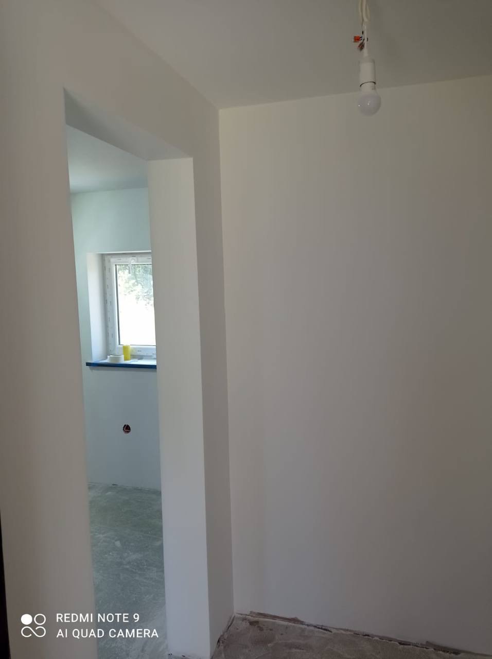 Wnętrze po malowaniu na biało, widoczny fragment przejścia do innego pomieszczenia z oknem, surowa żarówka na kablu, ślady po remoncie na podłodze.