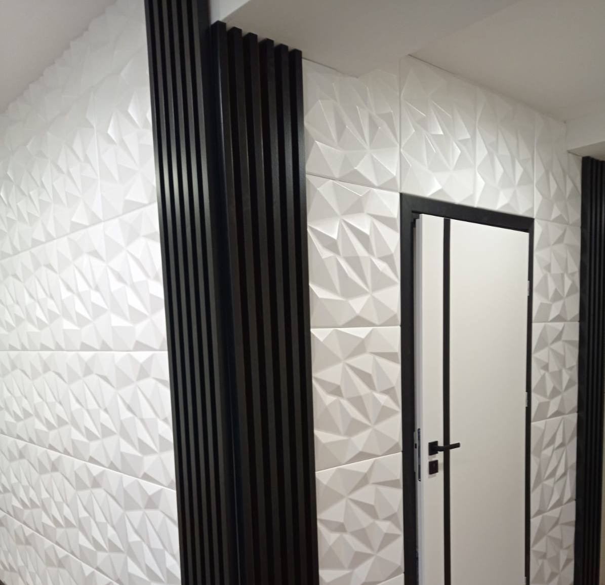 Ściana wykończona białymi trójwymiarowymi płytkami dekoracyjnymi, uzupełniona pionowymi, ciemnymi listwami drewnianymi i białymi drzwiami z czarną ramą.