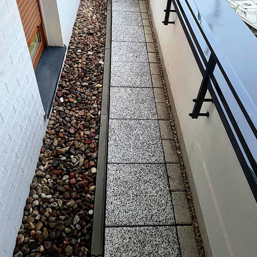 Wąski chodnik wyłożony szarymi płytami z drobnym wzorem, obok ściany z oknem i dekoracyjnym pasem z kamieni.