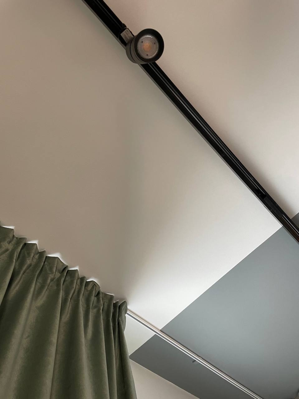 Fragment sufitu z szynoprzewodem oświetleniowym z reflektorem, pomalowanego na biało i szaro, z fragmentem zielonej zasłony w rogu.