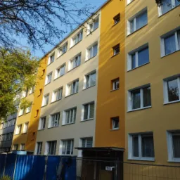 Wielopiętrowy budynek pomalowany na różne odcienie żółtego i beżu, z białymi ramami okiennymi, widoczny z poziomu ulicy, z fragmentem niebieskiego ogrodzenia i zieleni drzew.