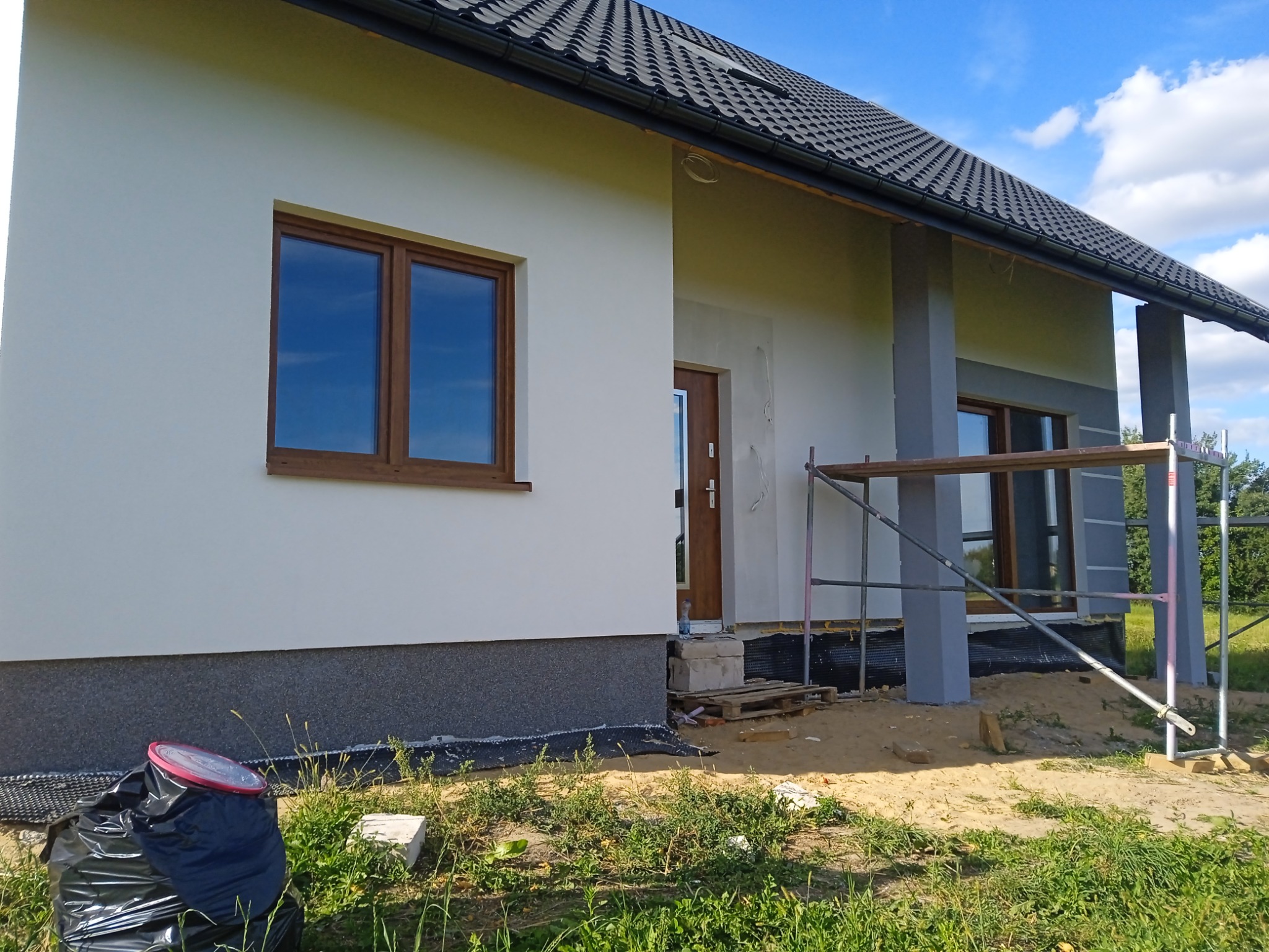 Nowo wybudowany dom jednorodzinny z szarą podmurówką i jasną elewacją, częściowo otynkowany, z widocznym rusztowaniem, drewnianą stolarką okienną i czarnym dachem z dachówki ceramicznej.