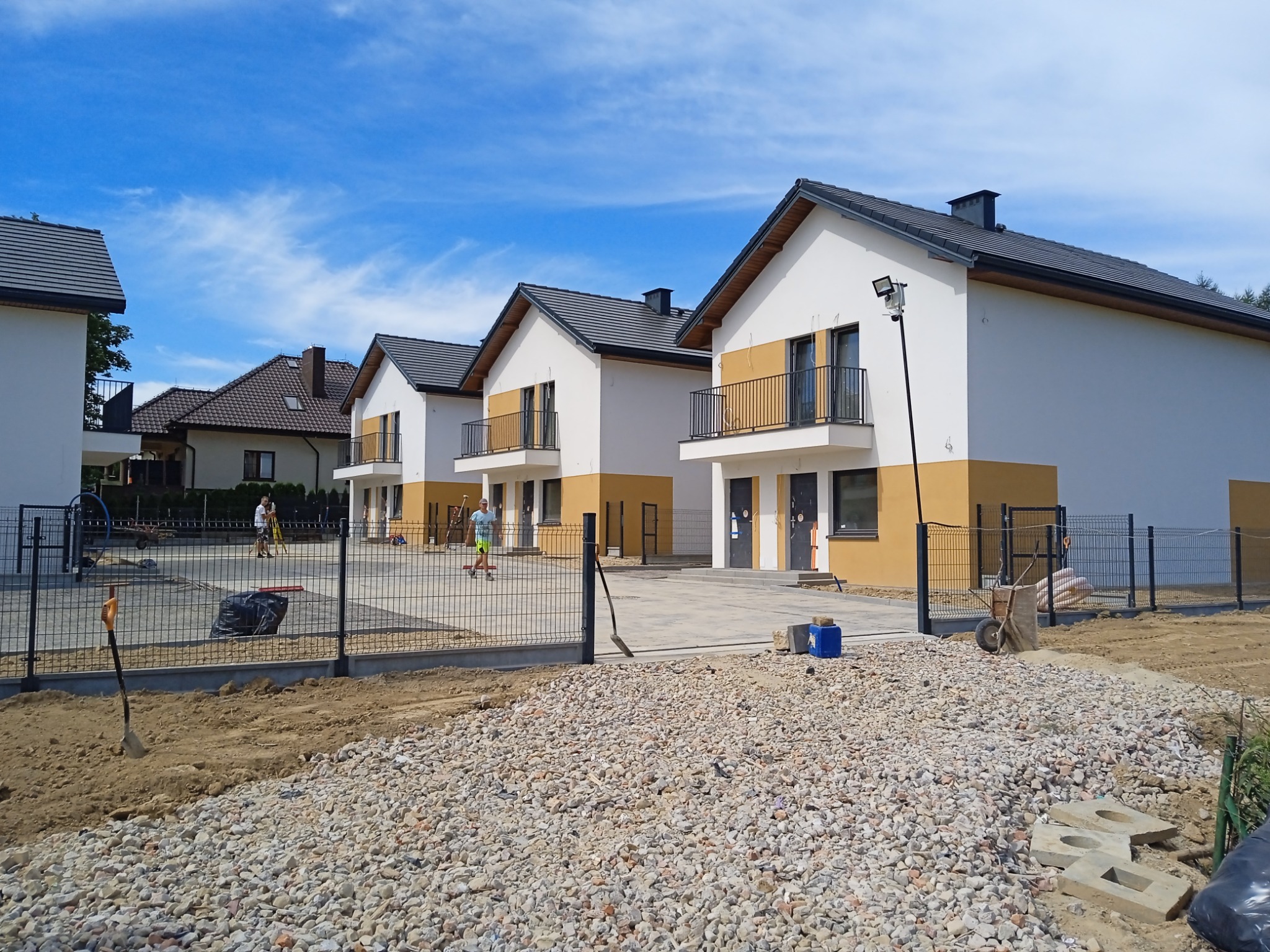 Nowe, szeregowe domy z białymi elewacjami i ciemnymi dachami, wykończone w części parterowej kolorem musztardowym, otoczone terenem przygotowanym pod zagospodarowanie, widoczne narzędzia i gruz.