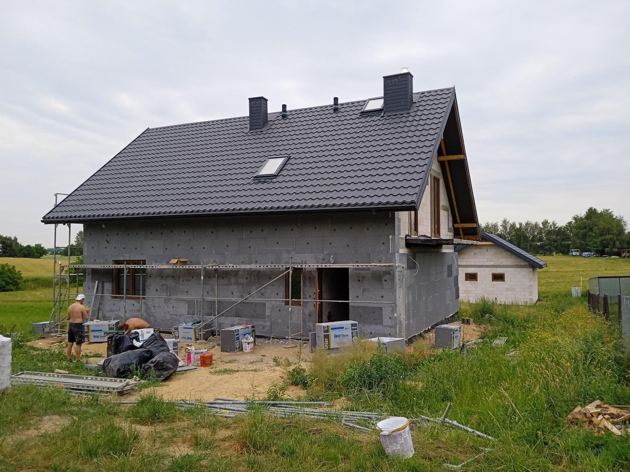 Dom w trakcie prac związanych z izolacją ścian zewnętrznych, widoczne rusztowania, materiały izolacyjne i pracownicy w tle, dach pokryty ciemną dachówką.
