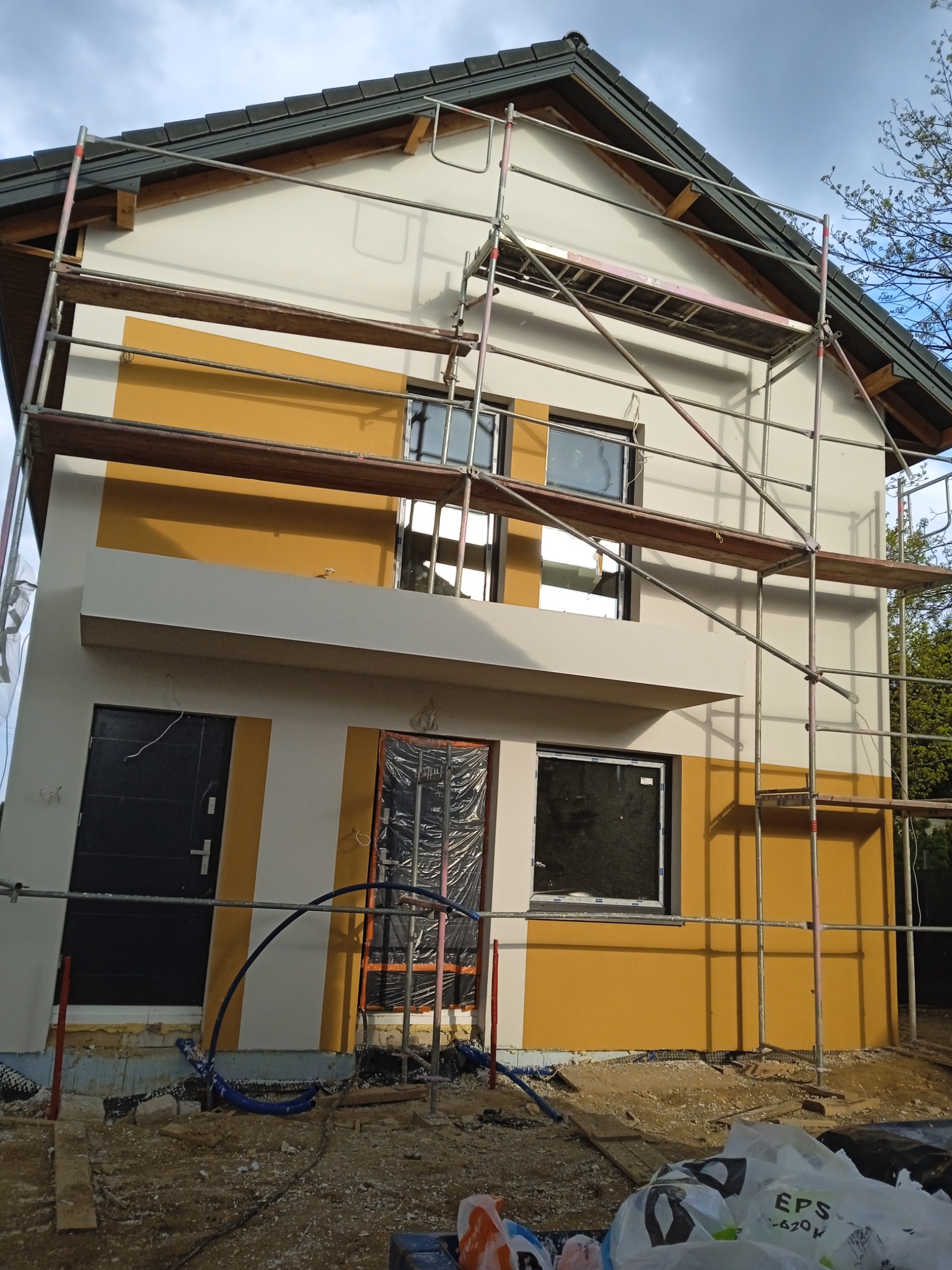 Ocieplanie elewacji domu jednorodzinnego z użyciem rusztowania, widoczne fragmenty izolacji fundamentów i przygotowane fragmenty elewacji w kolorze żółtym i białym.