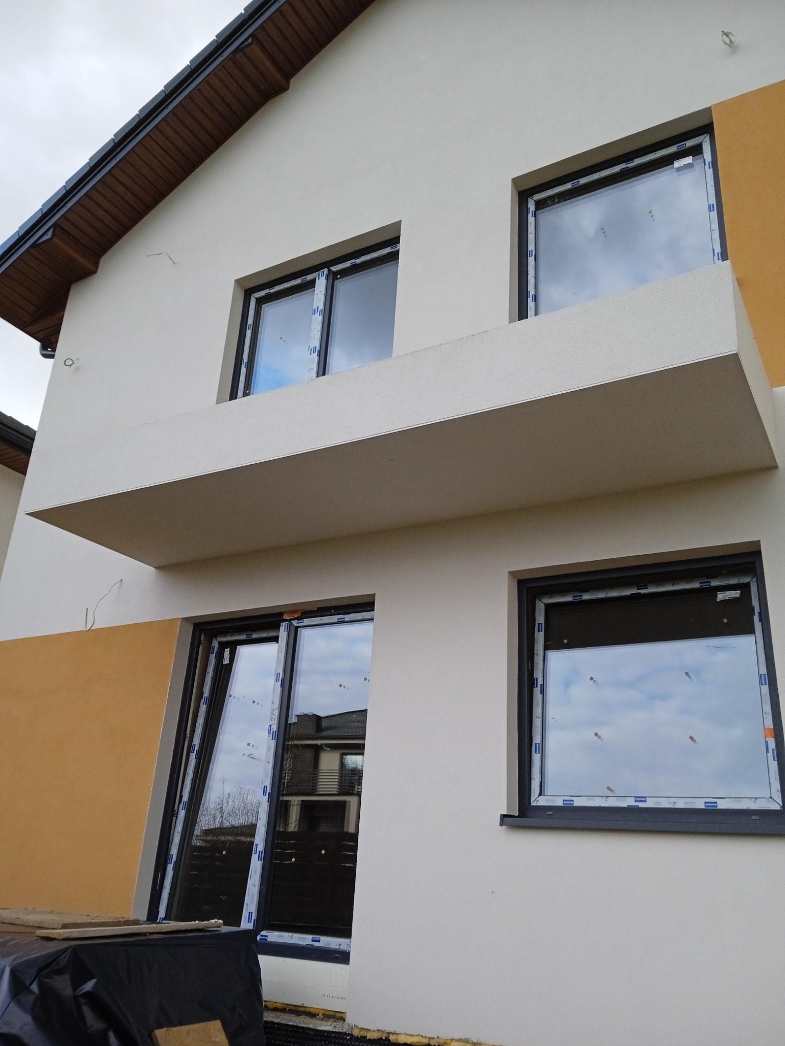 Dwukondygnacyjny dom z jasną elewacją, balkonem i oknami z czarnymi ramami, częściowo wykończony, z widocznym ociepleniem przy gruncie i fragmentem żółtej ściany.