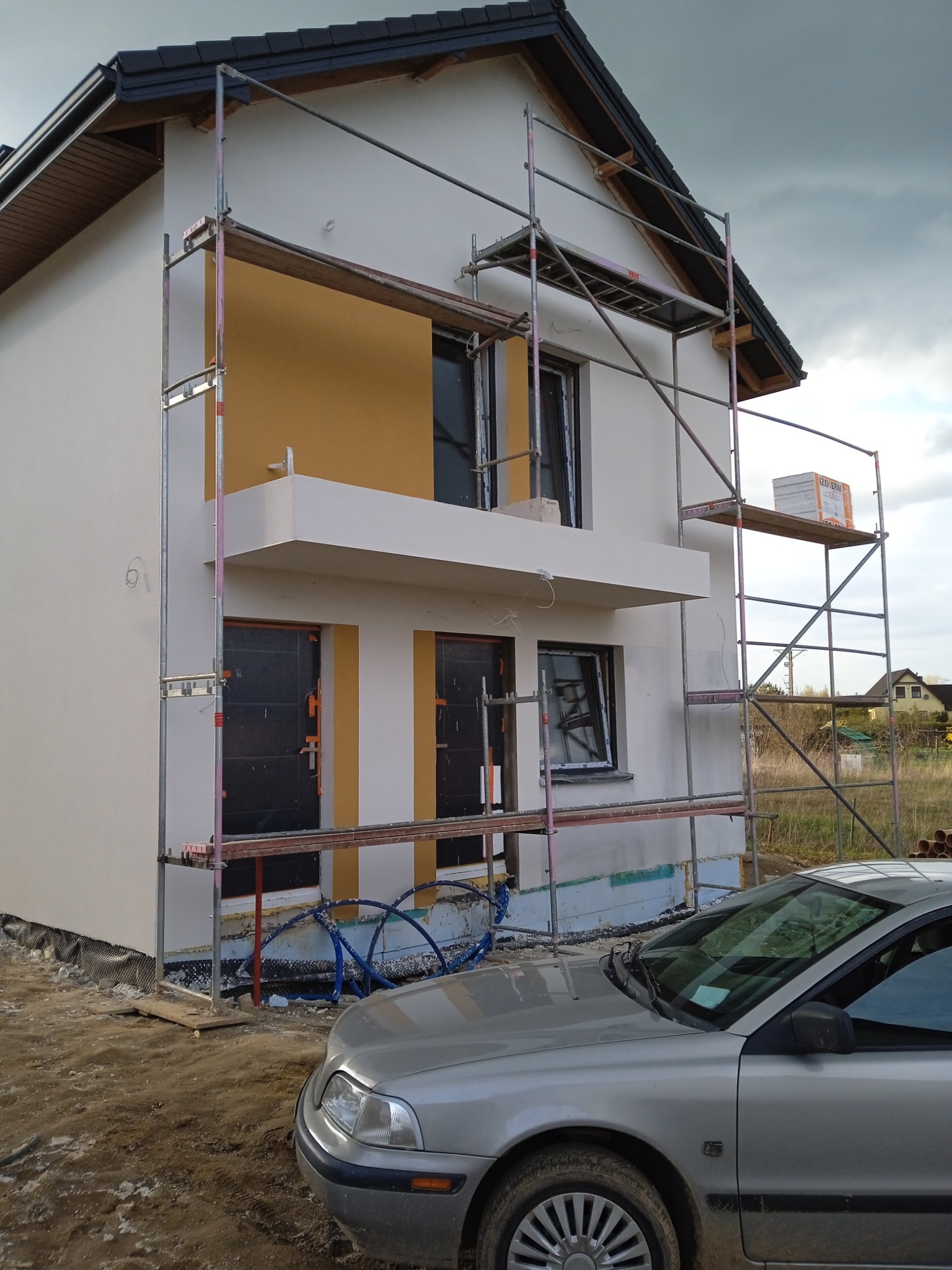 Dwukondygnacyjny dom jednorodzinny w trakcie prac elewacyjnych, widoczne rusztowania, częściowo otynkowana elewacja w kolorze kremowym z fragmentem w kolorze musztardowym, okna zabezpieczone folią,...