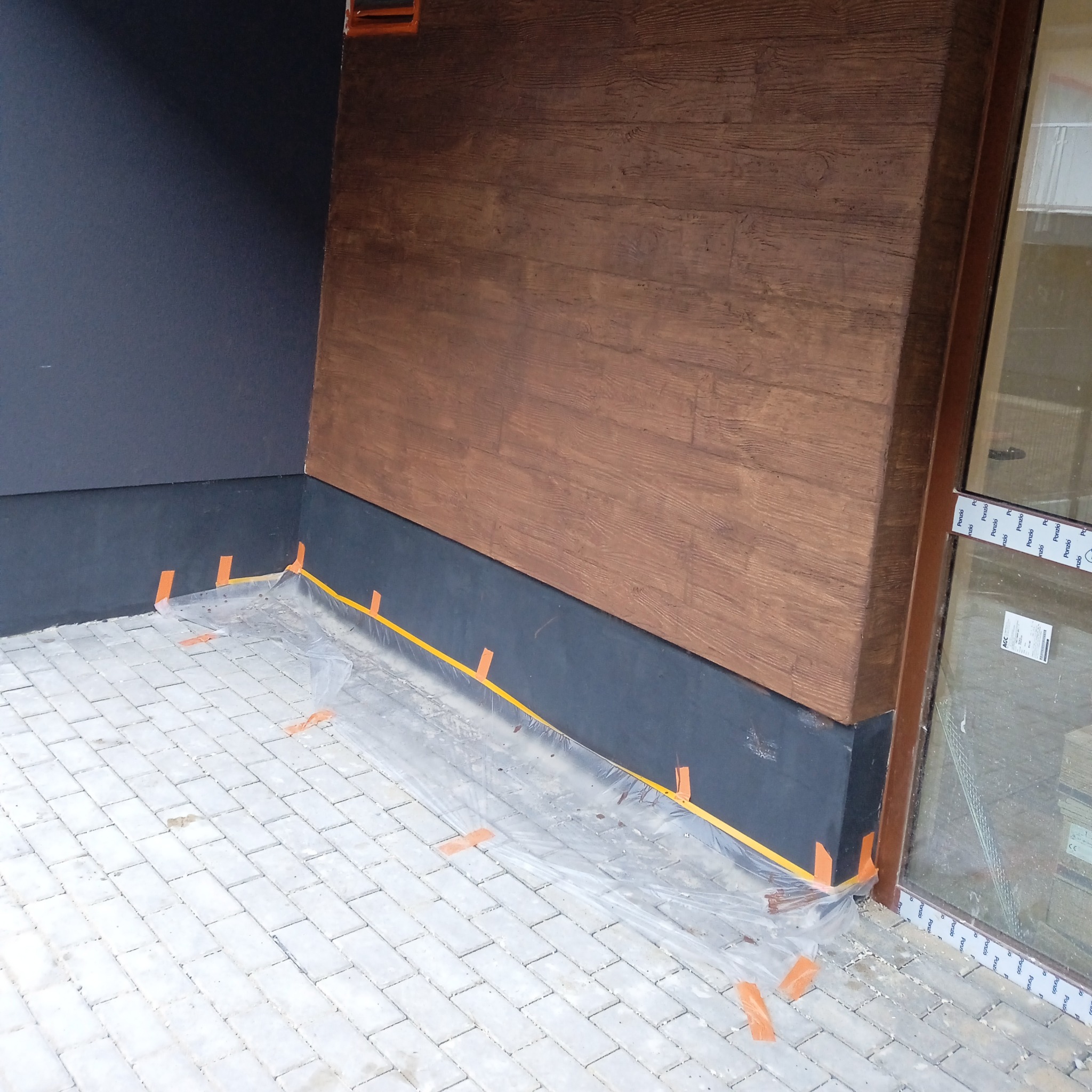 Fragment elewacji budynku z wykończeniem imitującym drewno, zabezpieczony folią i taśmą malarską na styku z chodnikiem z kostki brukowej podczas prac wykończeniowych.