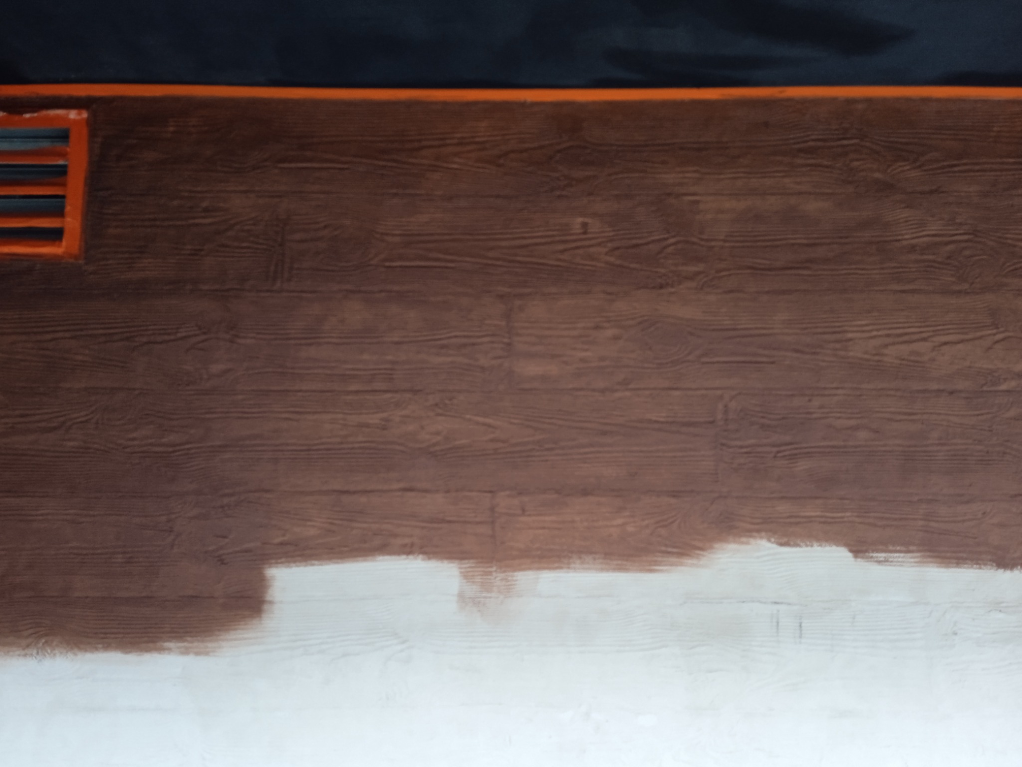 Fragment brązowej elewacji imitującej drewno, częściowo pomalowanej na biało, z pomarańczową kratką wentylacyjną w lewym górnym rogu.