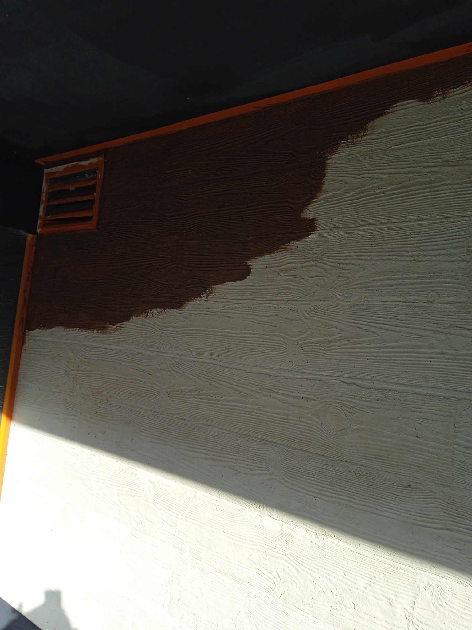 Fragment ściany zewnętrznej z fakturą drewna, częściowo pomalowanej na brązowo, z kratką wentylacyjną w rogu, w trakcie prac wykończeniowych.