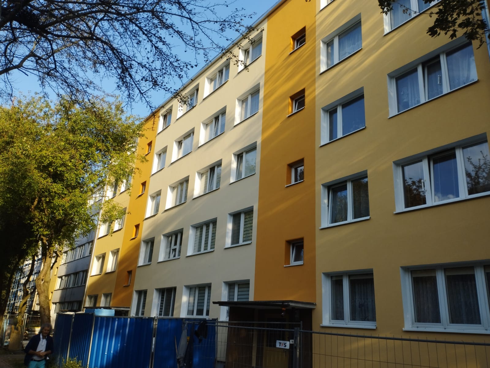 Wielopiętrowy budynek pomalowany na różne odcienie żółtego i beżu, z białymi ramami okiennymi, widoczny z poziomu ulicy, z fragmentem niebieskiego ogrodzenia i zieleni drzew.