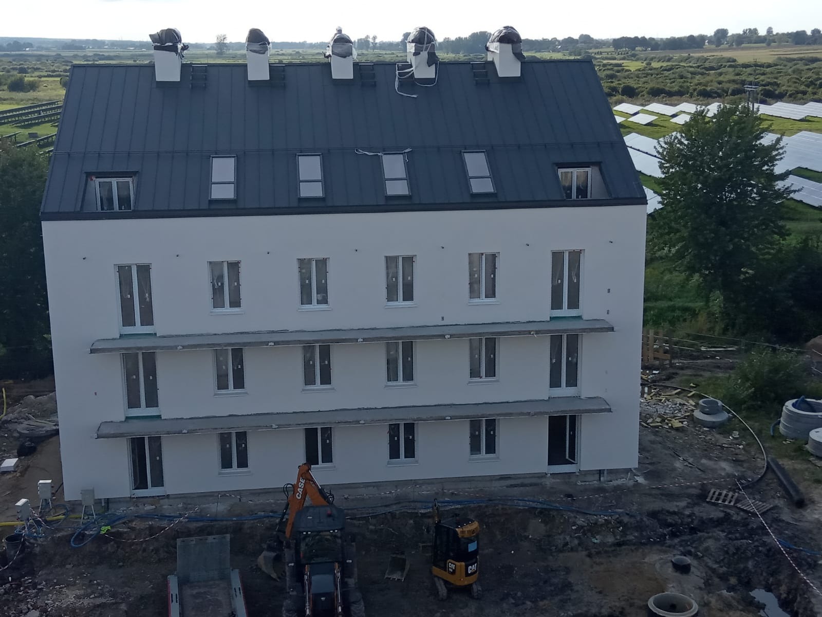 Trzypiętrowy budynek mieszkalny z nową, jasną elewacją i ciemnym dachem, widoczny z góry, z dwoma małymi koparkami na placu budowy i panelami słonecznymi w tle.