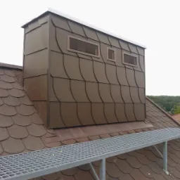 Dach z brązową dachówką karpiówką i obudowany komin z oknami, zabezpieczony metalową platformą. Widok z góry na tle zieleni.