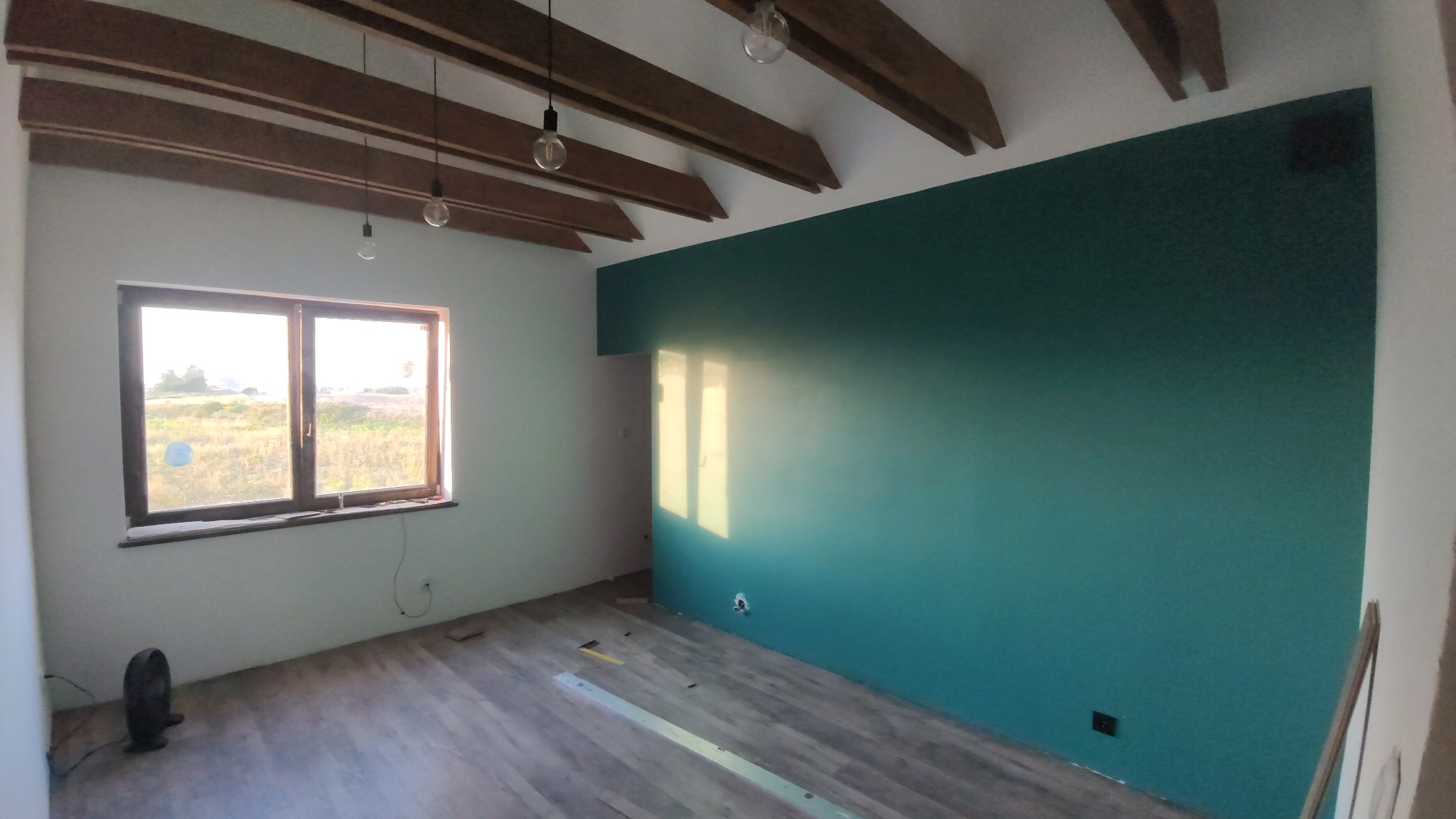 Wnętrze pokoju po malowaniu ścian na zielono, z drewnianym stropem, oświetleniem wiszącym i oknem z widokiem na zewnątrz. Podłoga drewniana, widoczne ślady po remoncie.