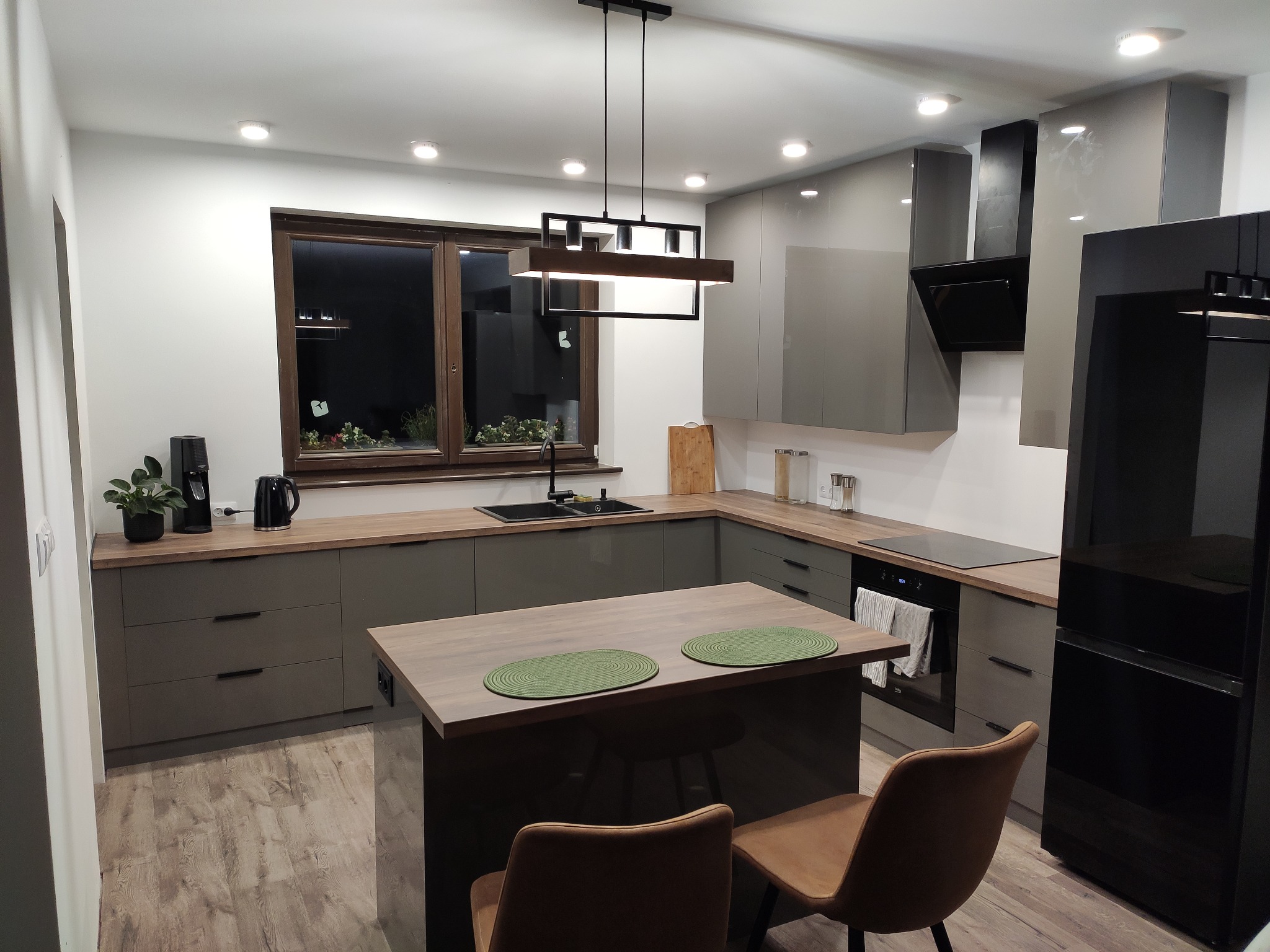 Nowoczesna kuchnia w szarościach z drewnianymi blatami, wyspą i minimalistycznym oświetleniem, widok z perspektywy wejścia, z dwoma krzesłami przy wyspie.