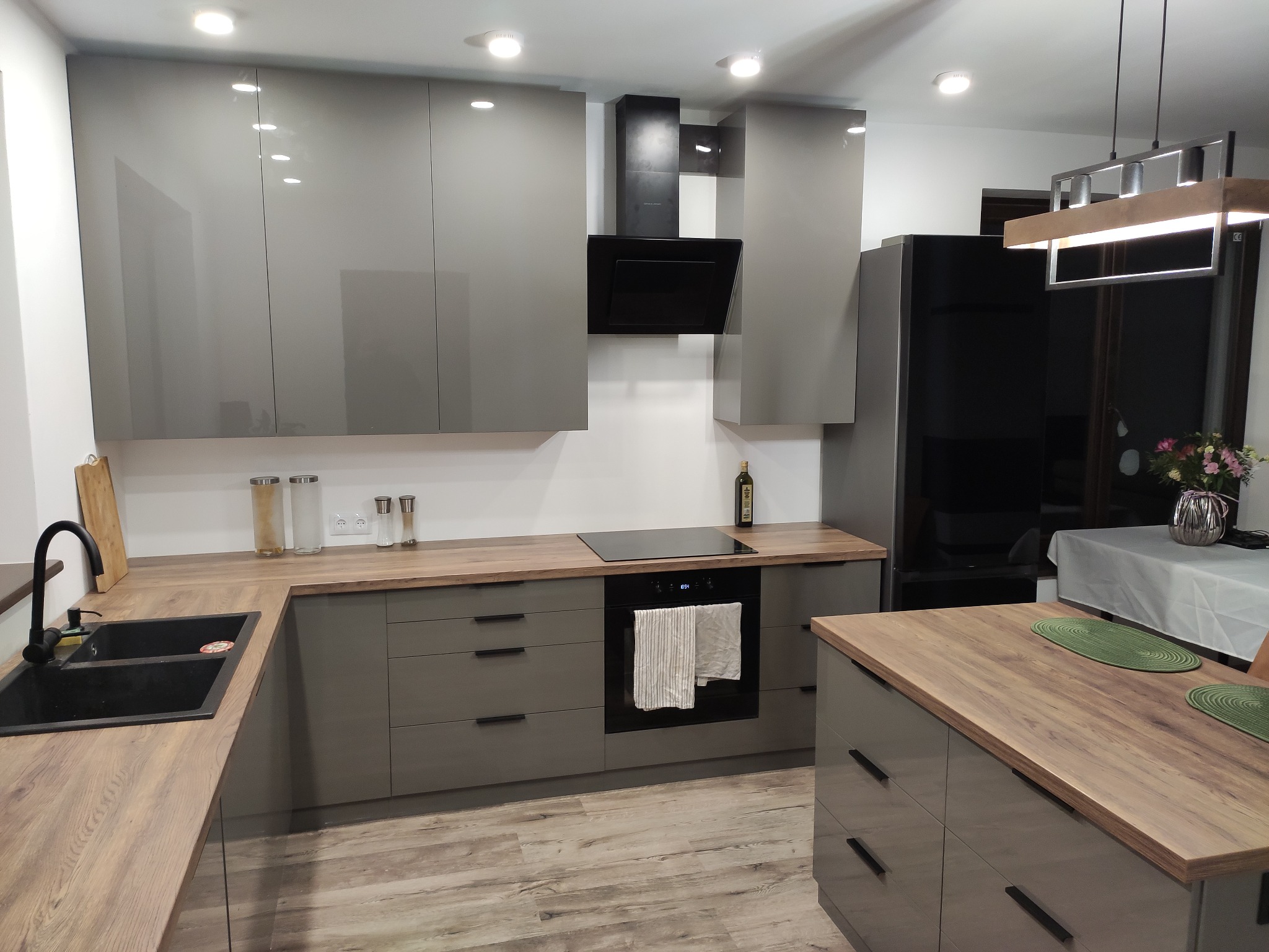Nowoczesna kuchnia w odcieniach szarości z drewnianymi blatami, czarnym zlewem i płytą indukcyjną; widoczny okap i oświetlenie sufitowe.