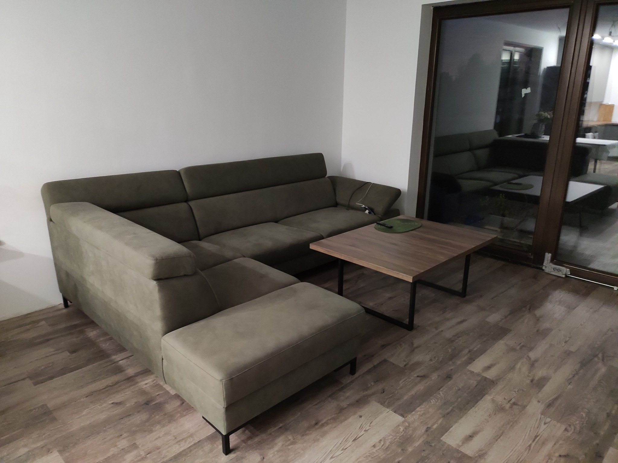 Szary narożnik z funkcją relaks w salonie z drewnianą podłogą i stolikiem kawowym na metalowych nogach, widok przez przesuwne okno na identyczne wnętrze.